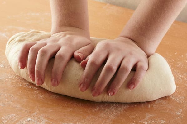 Two hands kneading a ball of dough on a floured wooden surface.