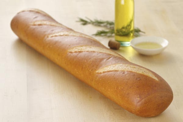 A freshly baked baguette with a golden-brown crust rests on a light wooden surface. Behind it, a bottle of olive oil, a small bowl of oil, and a sprig of rosemary are placed, suggesting a simple, rustic setting.