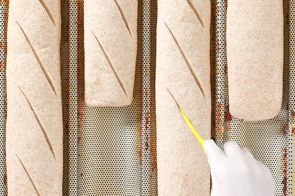 A gloved hand uses a yellow knife to score diagonal lines on several unbaked baguettes arranged on a perforated baking sheet. The dough has a light, floury appearance.