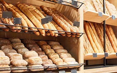 A bakery display featuring an assortment of breads. The top shelves hold long baguettes, while the middle shelves house a variety of rolls, buns, and loaves. The breads are neatly arranged in a well-lit wooden shelving unit.