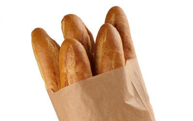 A paper bag holds four freshly baked baguettes. The baguettes have a golden-brown crust and are partially visible above the top of the bag, set against a plain white background.