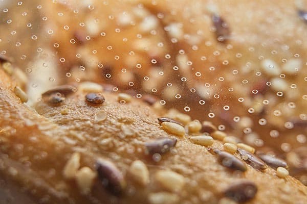 Close-up image of a bread crust topped with various seeds, including sesame and flax seeds. The surface is dotted with condensation droplets, creating a textured and detailed appearance of the freshly baked bread.