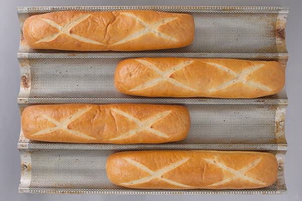 Four freshly baked baguettes, each with a scored X pattern on top, are laid out on a textured metal baking tray. The bread has a golden-brown crust and is aligned in parallel rows. The tray is slightly worn, indicating frequent use.