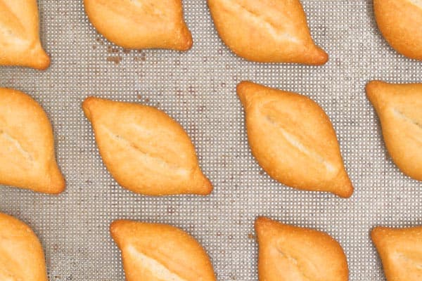 Four freshly baked baguettes, each with a scored X pattern on top, are laid out on a textured metal baking tray. The bread has a golden-brown crust and is aligned in parallel rows. The tray is slightly worn, indicating frequent use.